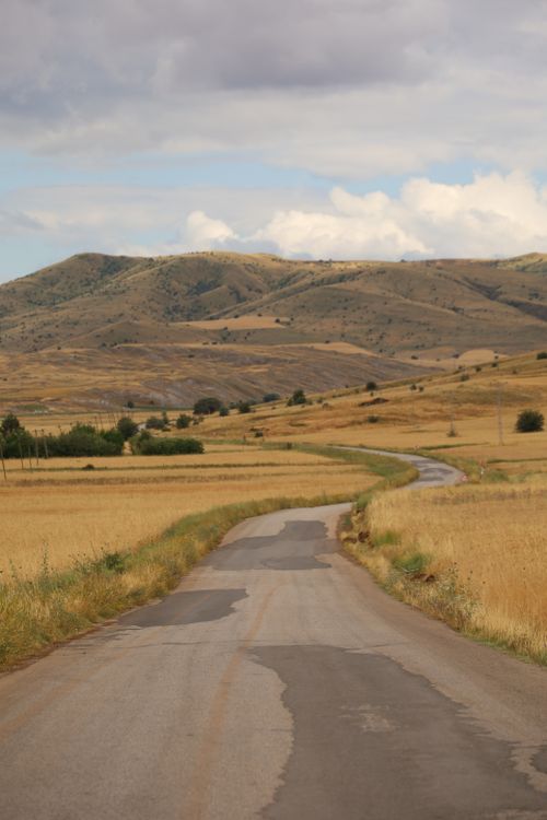 Free A scenic winding road through golden fields and rolling hills under a cloudy sky. Stock Photo