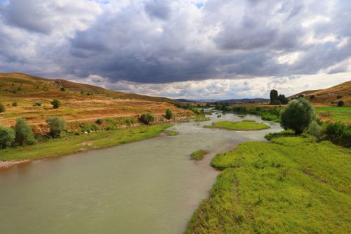 Free A peaceful river flowing through lush fields under dramatic cloudy skies. Stock Photo