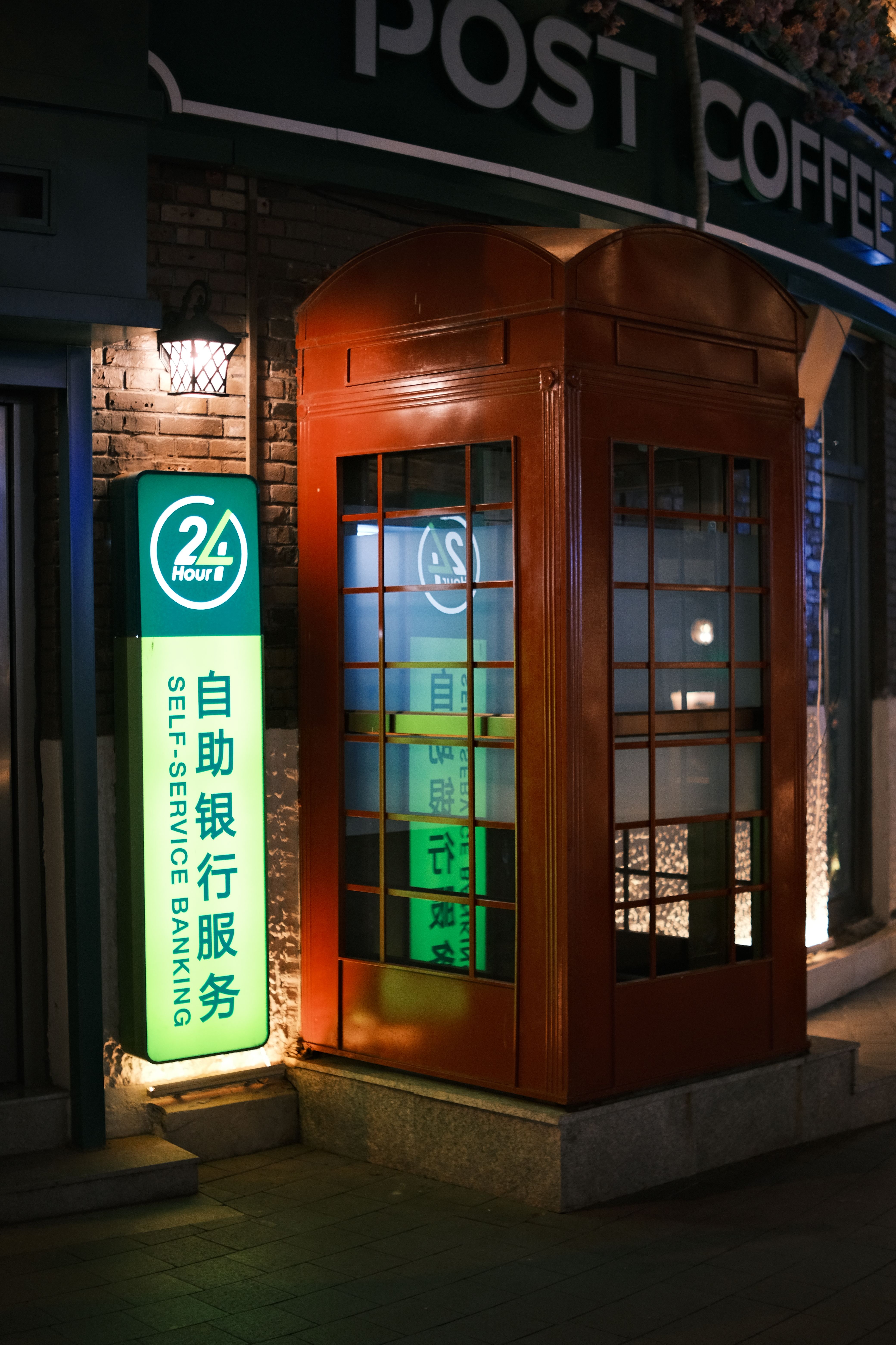 Night view of a bright red telephone-style booth beside a self-service banking sign in an urban setting.