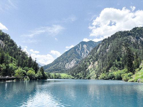 Free Breathtaking mountain lake view surrounded by lush greenery under a clear blue sky. Stock Photo
