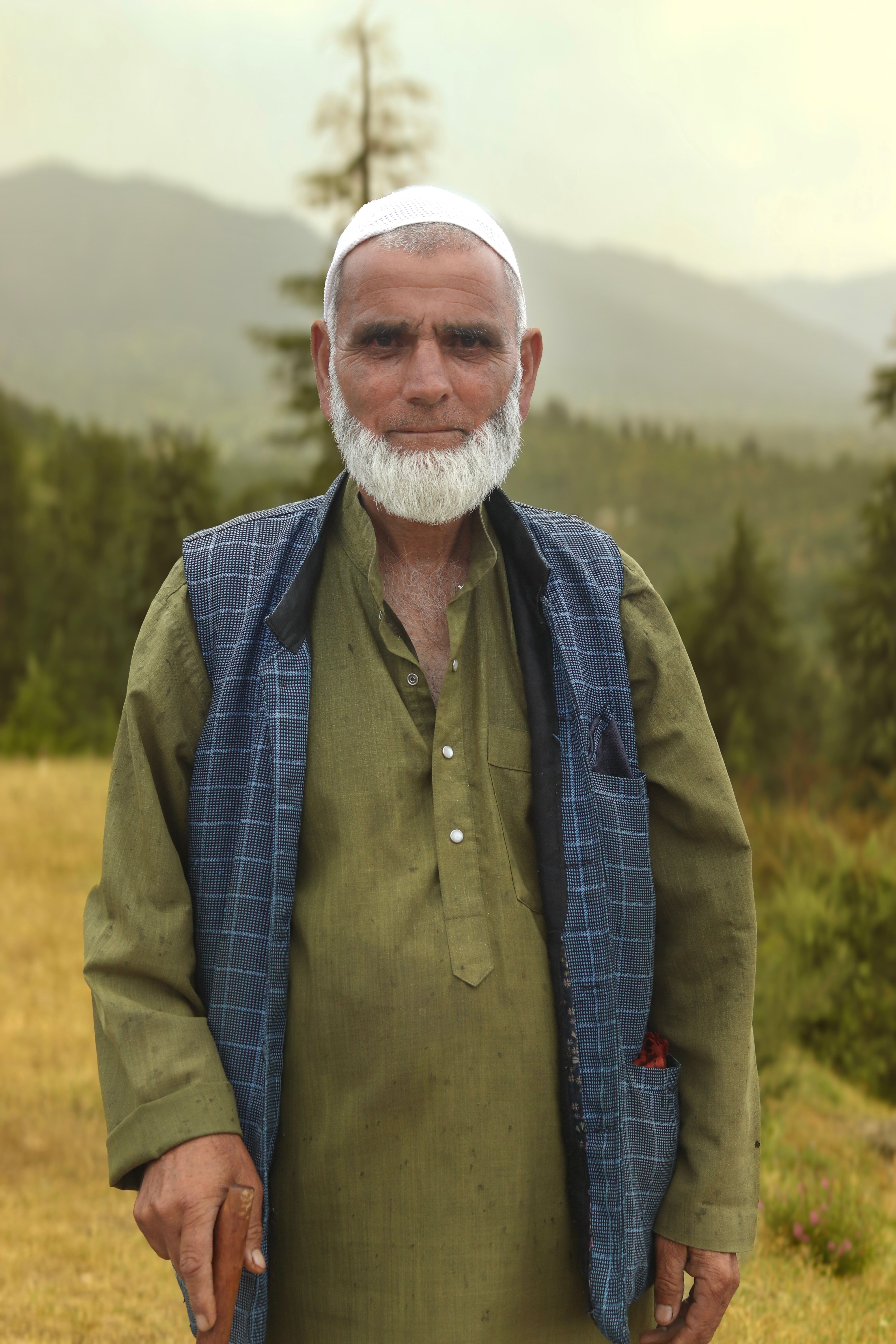 Portrait of an elderly man in traditional attire, standing in a mountainous landscape.