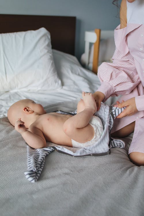 Free Side view of adorable little baby in diaper lying on comfortable bed in light bedroom while crop unrecognizable mother dressing newborn in striped suit at home Stock Photo