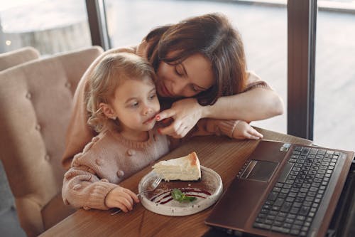 Careful young woman in casual clothes sitting at table with laptop and wiping girls mouth while daughter sitting near mother eating yummy cake and looking at screen in cafe