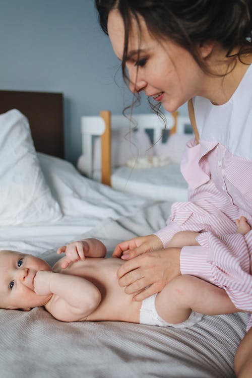 Woman in White and Pink Striped Long Sleeve Shirt Playing with Baby Lying on Bed