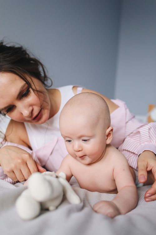 Free Calm baby lying on bed with mother and looking at toy while resting in bedroom during weekend morning at home Stock Photo