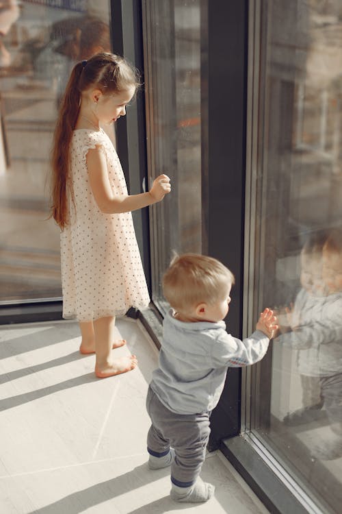 Side view of cheerful children wearing casual clothes standing near window and looking at reflection while having rest in modern house