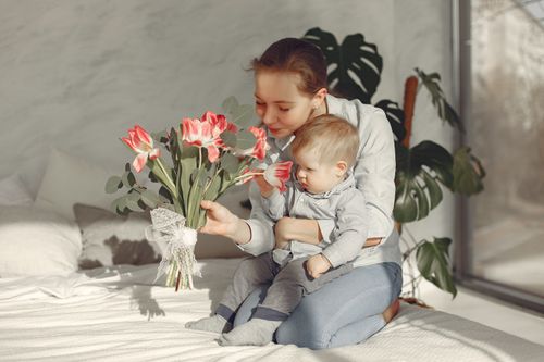 Free Cheerful mother smiling while playing and sitting with toddler on bed and spending weekend day together in home looking at flower Stock Photo