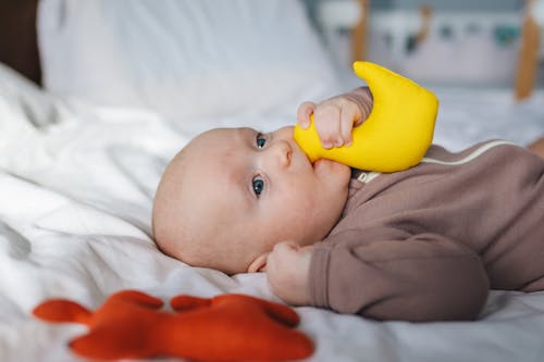 Side view of adorable newborn in brown wear lying on bed with clenched fist and yellow toy in mouth