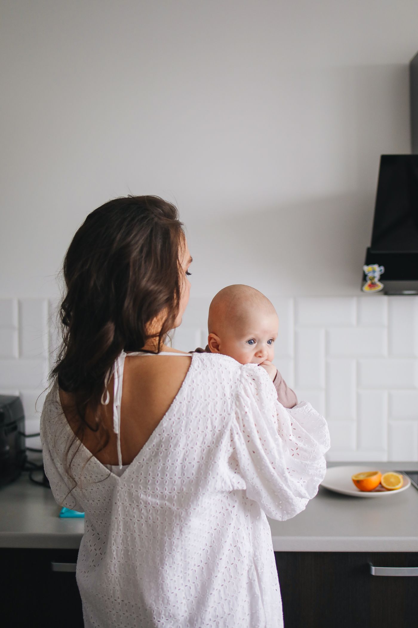Maman en robe blanche portant son bébé dans une cuisine moderne et lumineuse