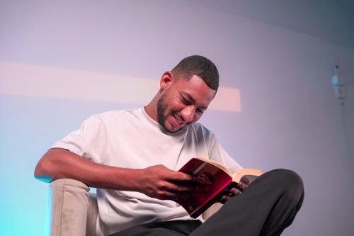 Free A smiling man sits on a chair, deeply engaged in reading a book. Stock Photo