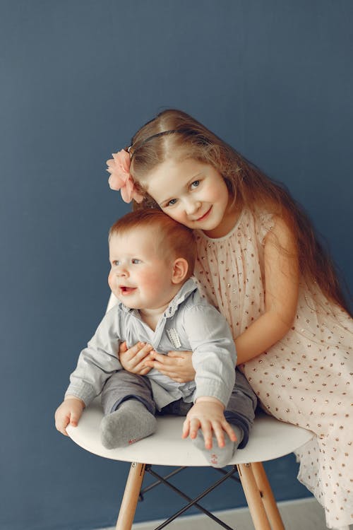 Happy red haired girl in polka dot dress looking at camera and hugging little brother while infant sitting on chair against gray background