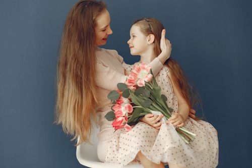 Free Side view of red haired lady in dress sitting on chair and cuddling cute girl with flowers while mom and daughter looking at each other Stock Photo
