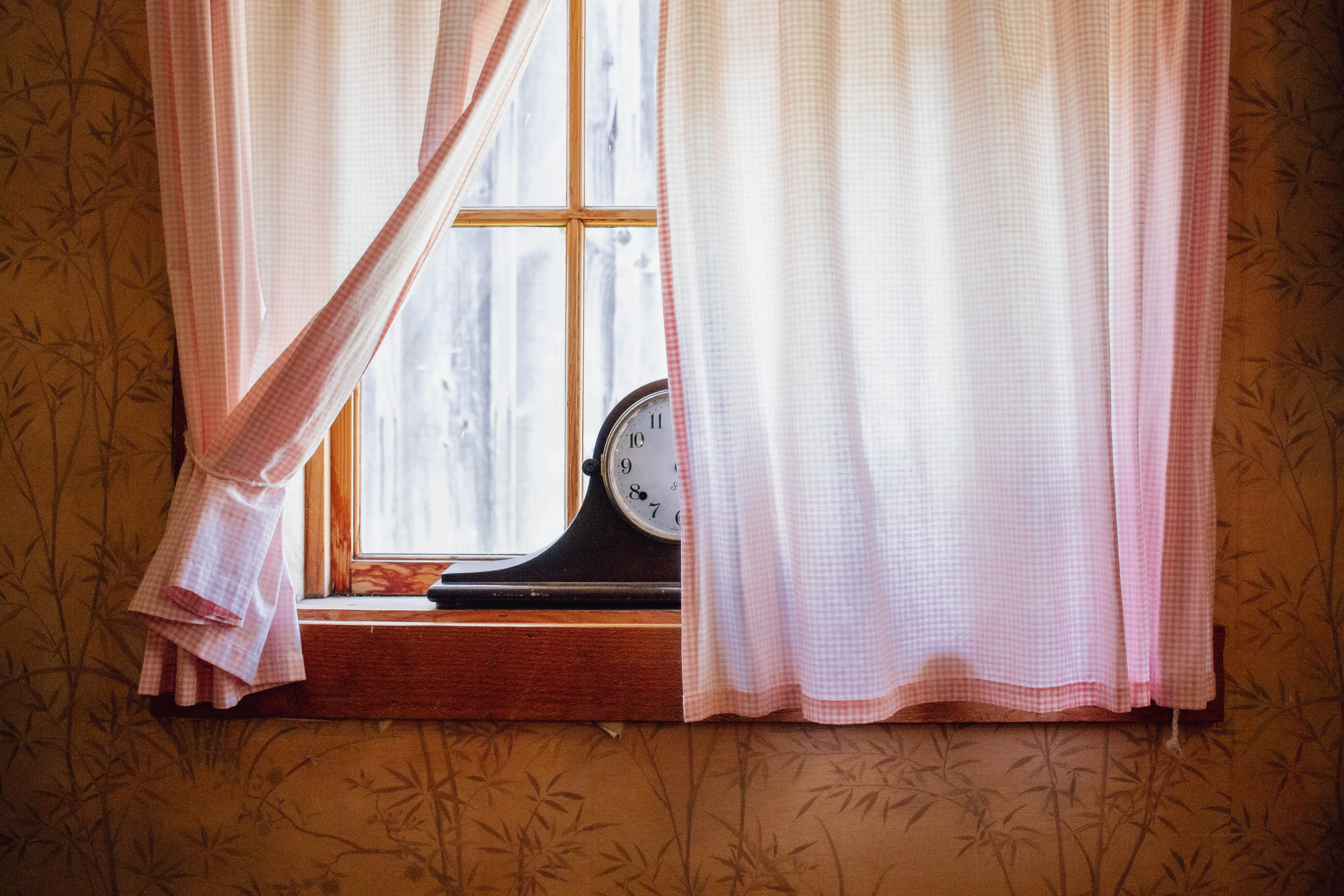 A vintage clock sits on a windowsill with pink checkered curtains and patterned wallpaper.