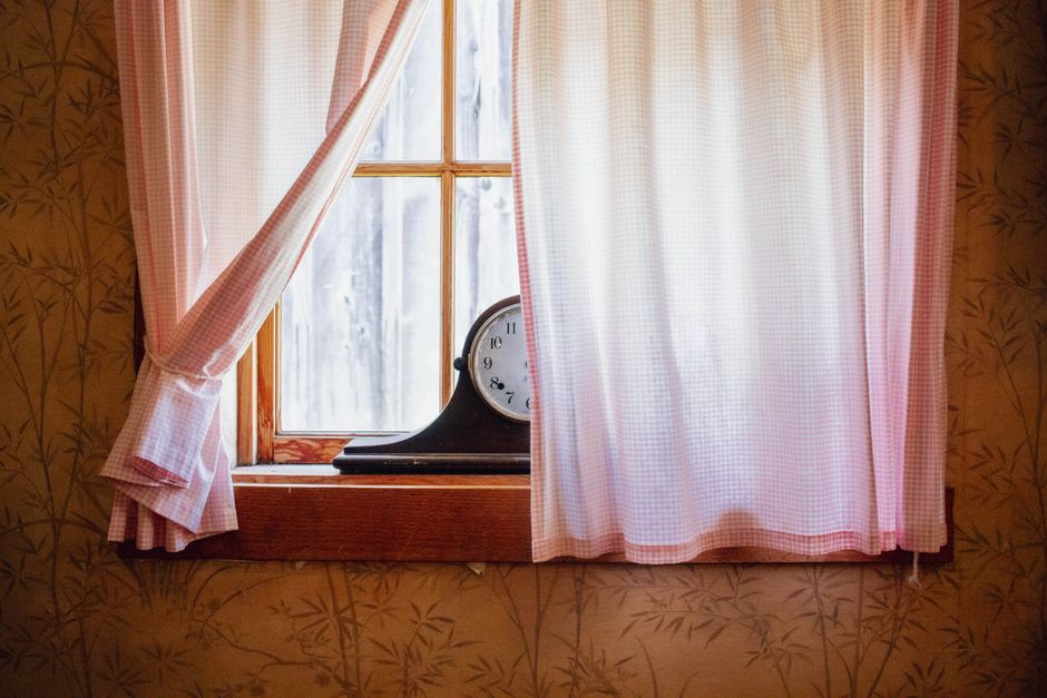 A vintage clock sits on a windowsill with pink checkered curtains and patterned wallpaper.