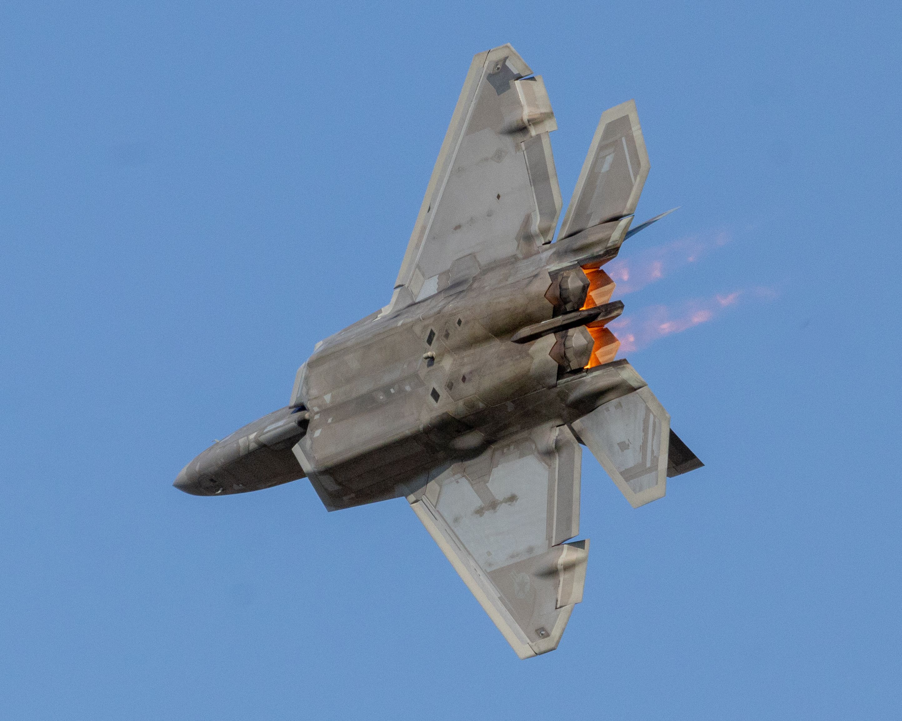 A fighter jet flying through a clear blue sky with glowing orange flames from its engines.