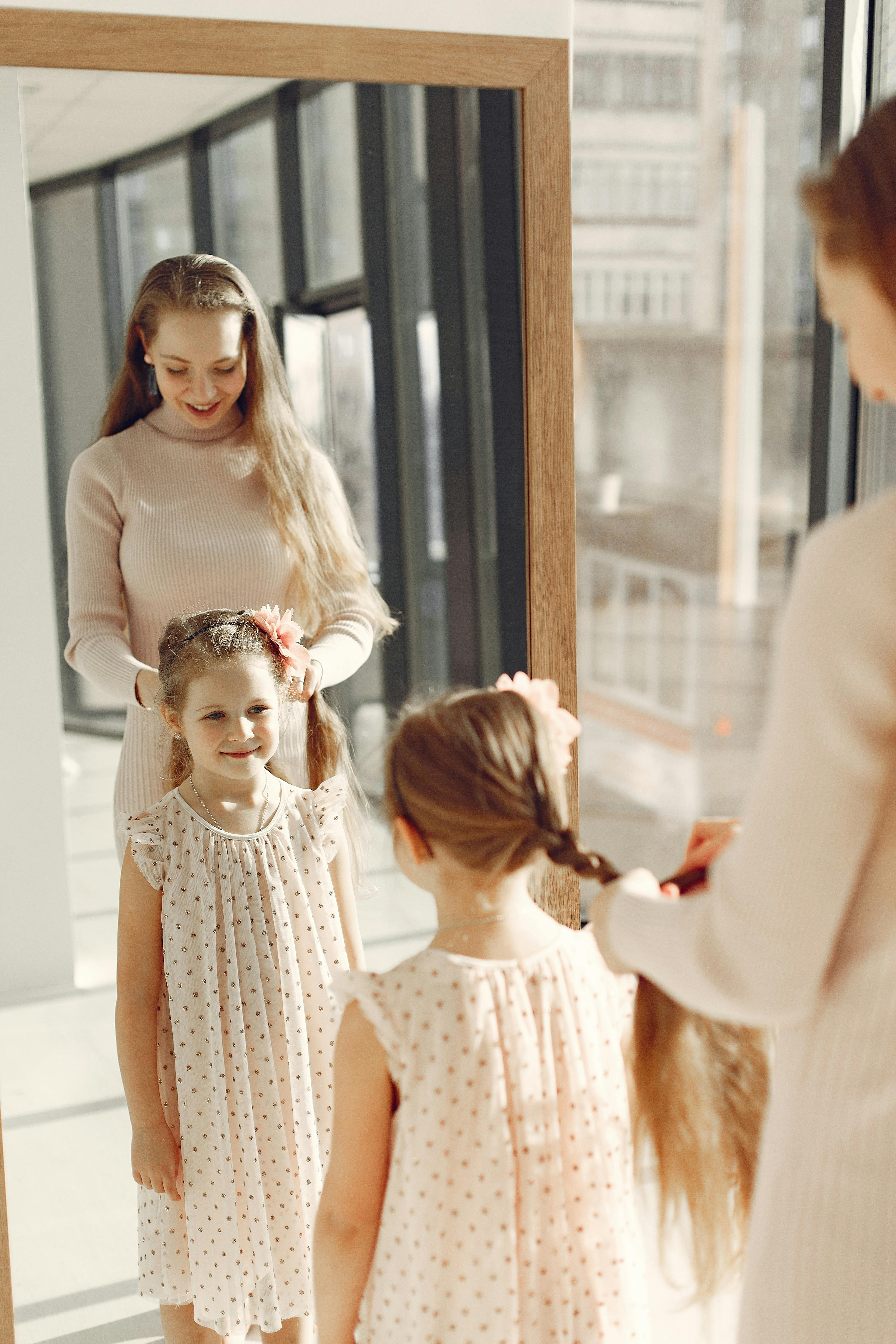 A mother braiding her smiling daughter's hair in front of a mirror in a sunlit room.