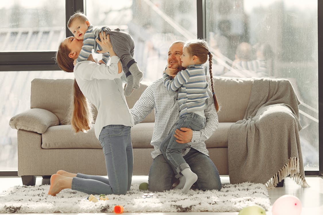 Free Cheerful mother kissing little boy while father hugging adorable daughter when family sitting on floor in rainy day against window Stock Photo