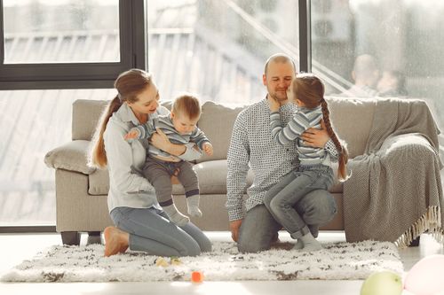Free Happy parents with children on knees in casual clothes sitting on floor and playing with kids while spending time together in living room near panoramic window Stock Photo