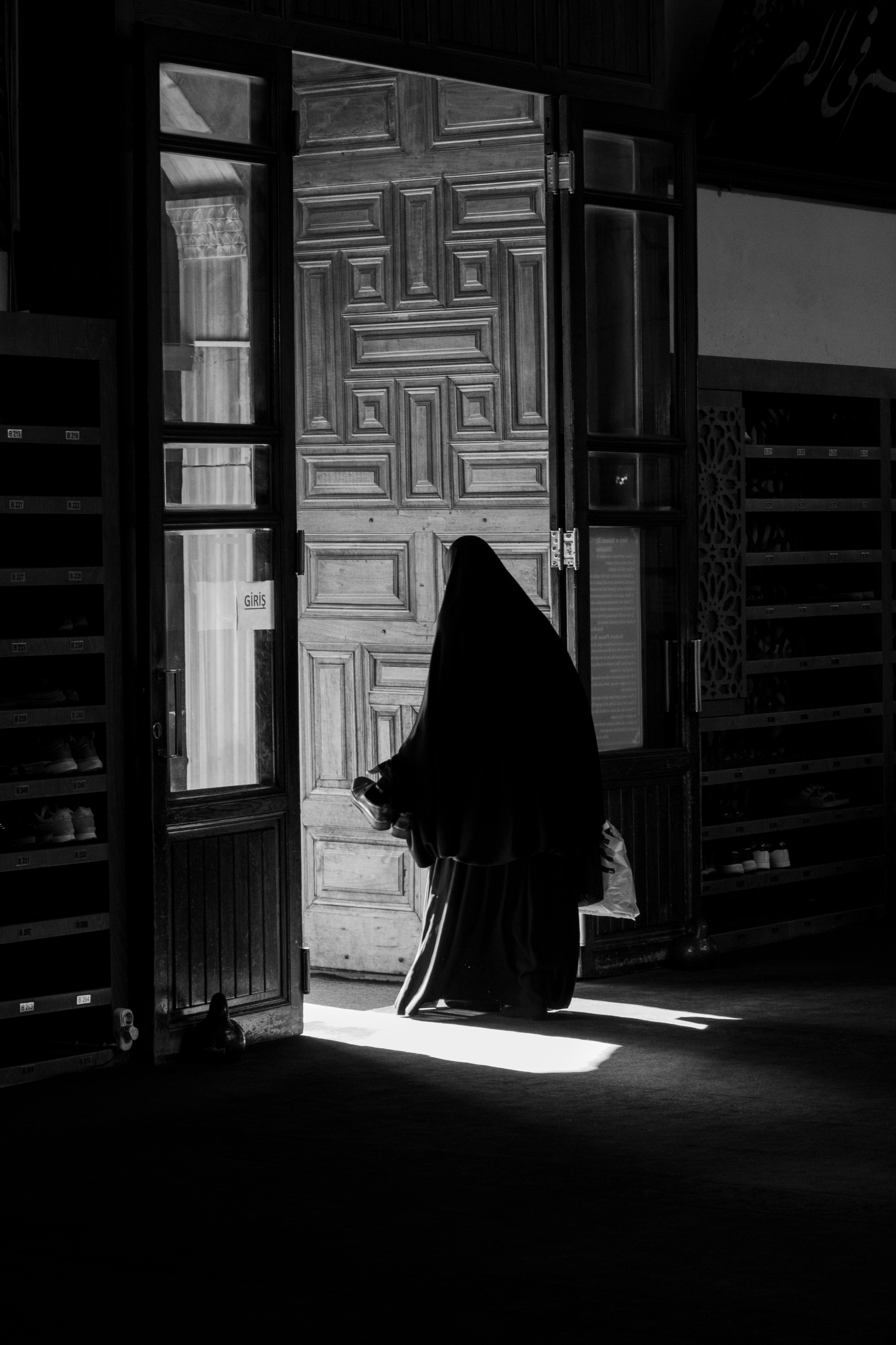 A woman in a traditional dress stands by a wooden door, captured in black and white.