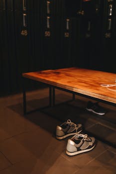 A pair of sneakers under a bench in a locker room with numbered lockers in the background.