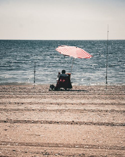 Free Man sitting on a beach under a striped umbrella by the ocean, enjoying a peaceful day. Stock Photo