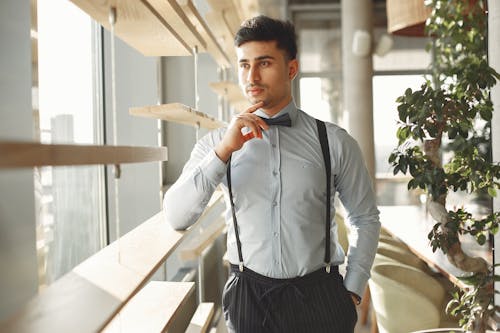 Pensive handsome ethnic male in elegant suit and bow tie standing in modern cafe with hand in pocket and looking through window