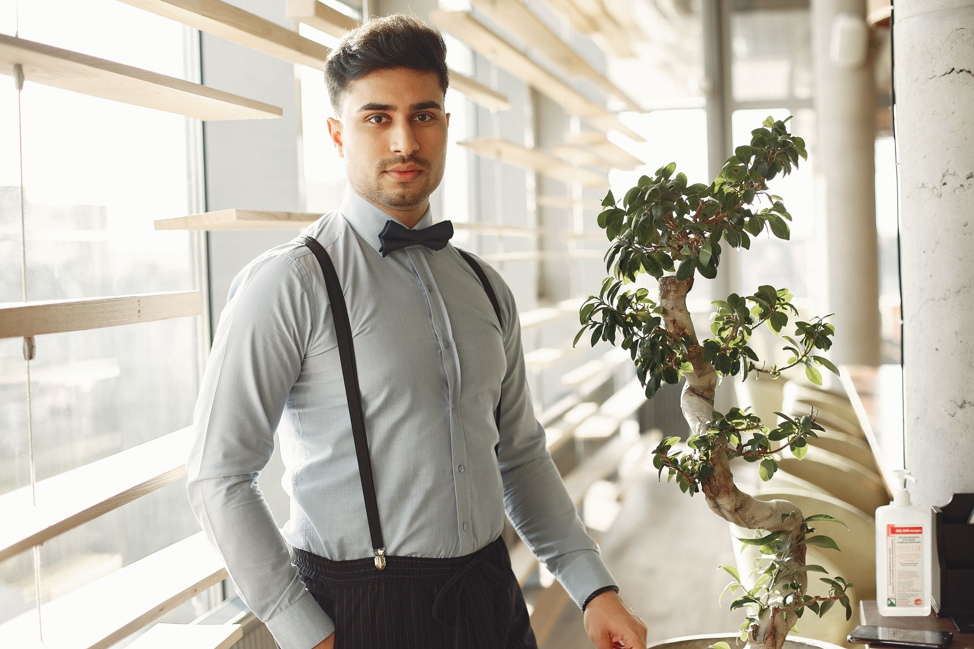 Portrait of a serious man in a bow tie standing indoors by a window with a bonsai tree.