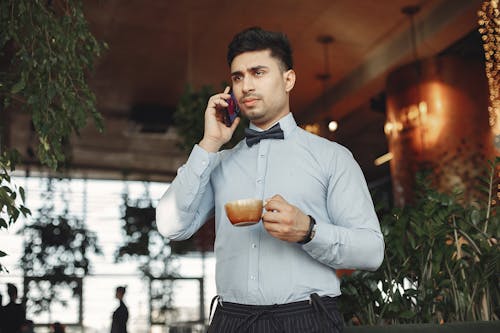 Busy handsome ethnic man in formal suit and bow tie standing in lobby of modern cafe with cup of hot coffee in hand and answering phone call while looking away