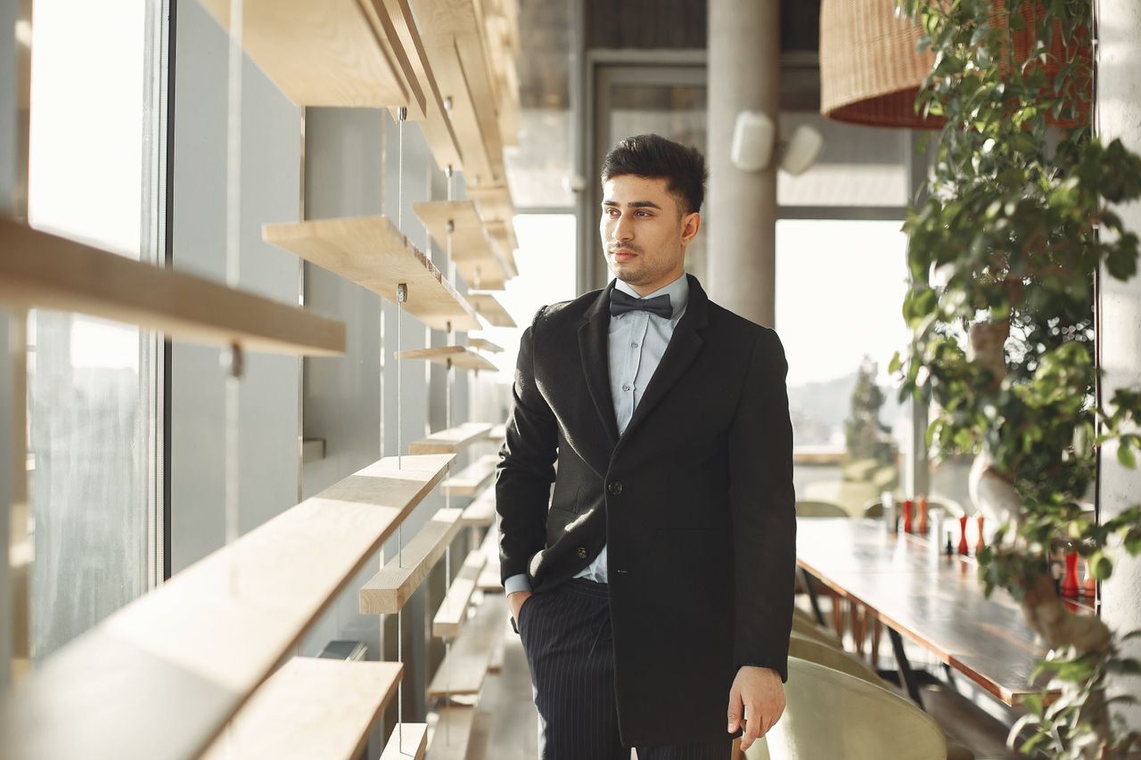 stylish man in a formal suit with a bow tie standing indoors by a sunl