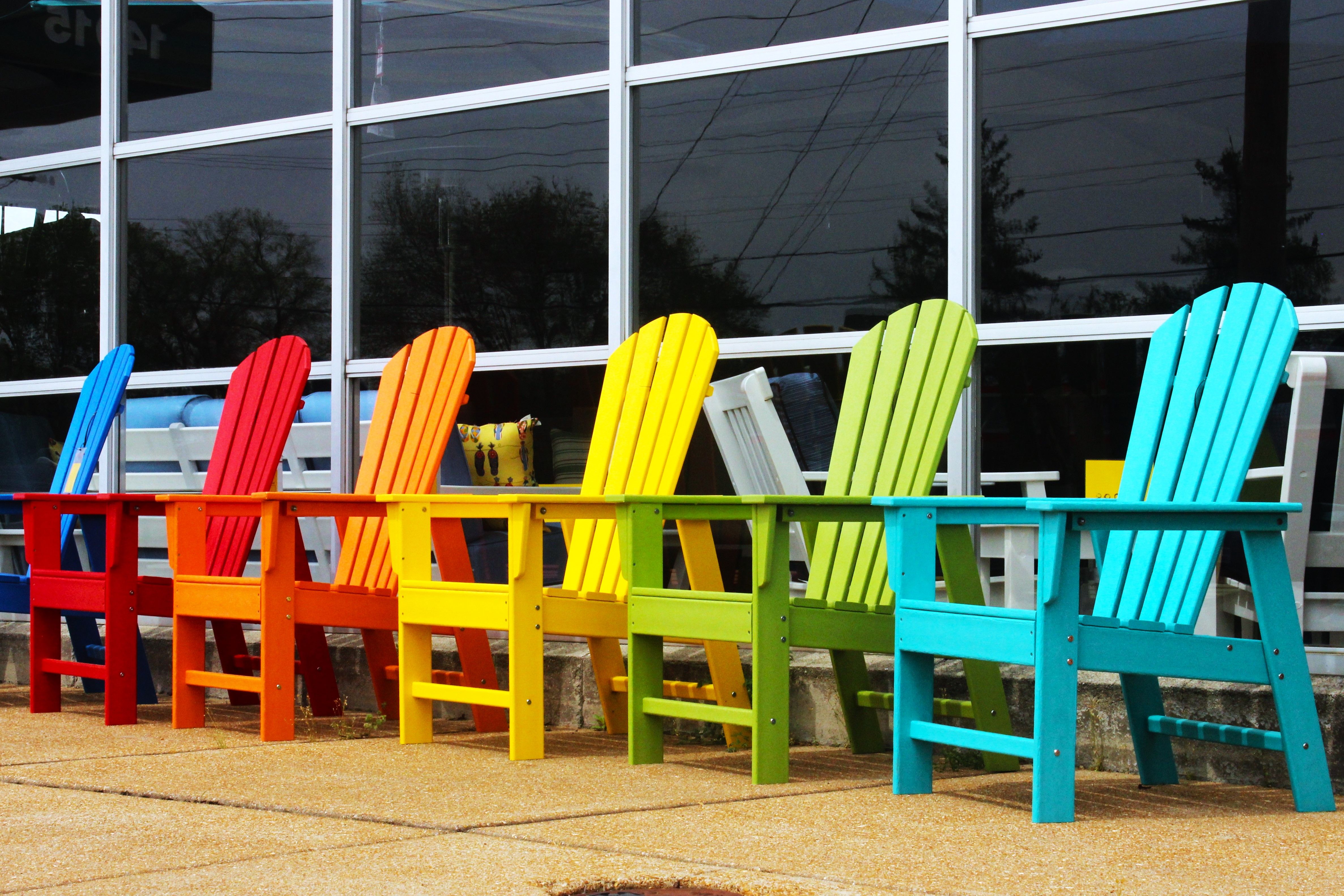 A vibrant row of Adirondack chairs in rainbow colors outside a glass building.