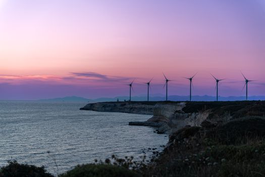 A serene view of wind turbines along a rocky coastline during a vibrant sunset.