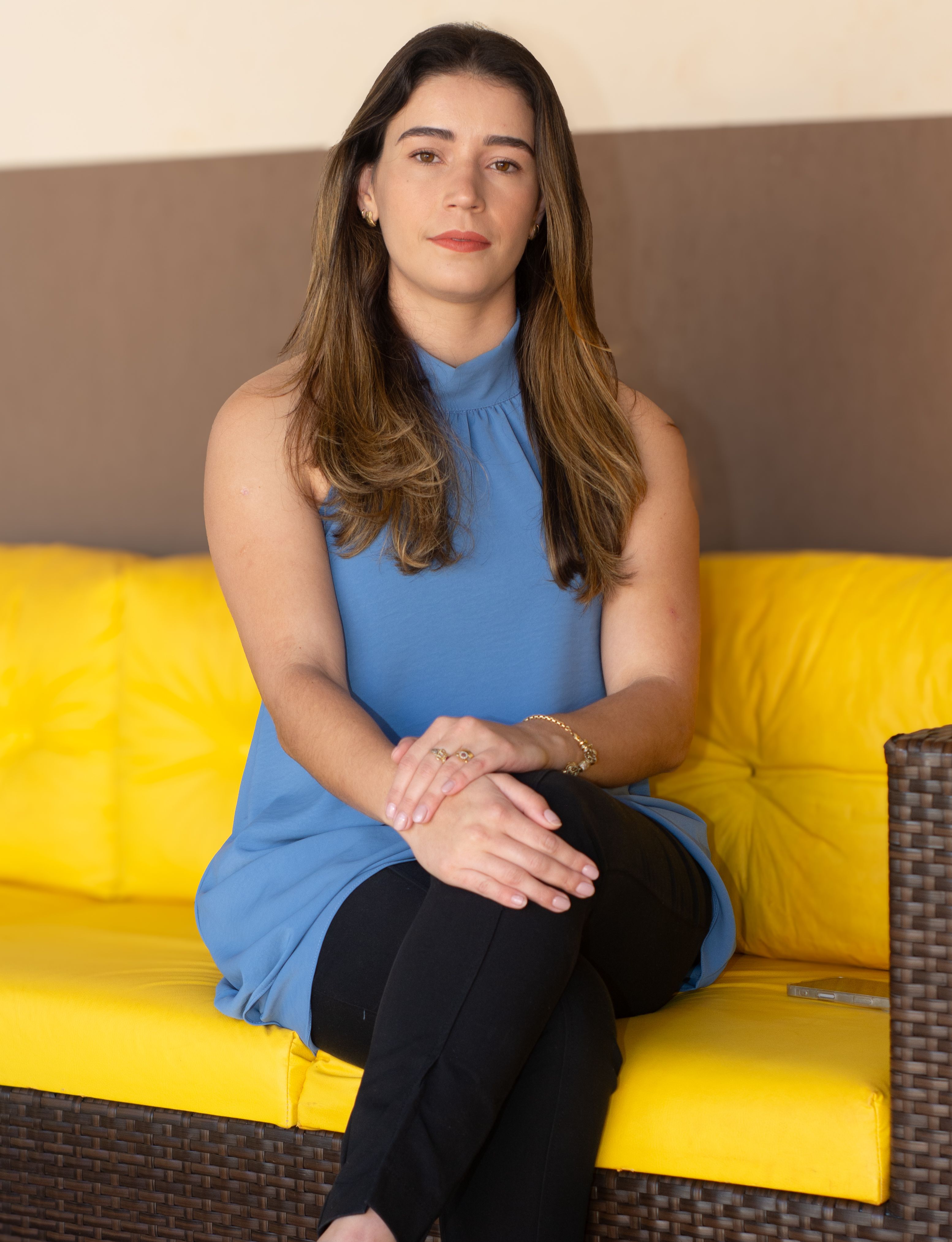 A young woman in a blue blouse and black pants sitting on a yellow couch indoors.