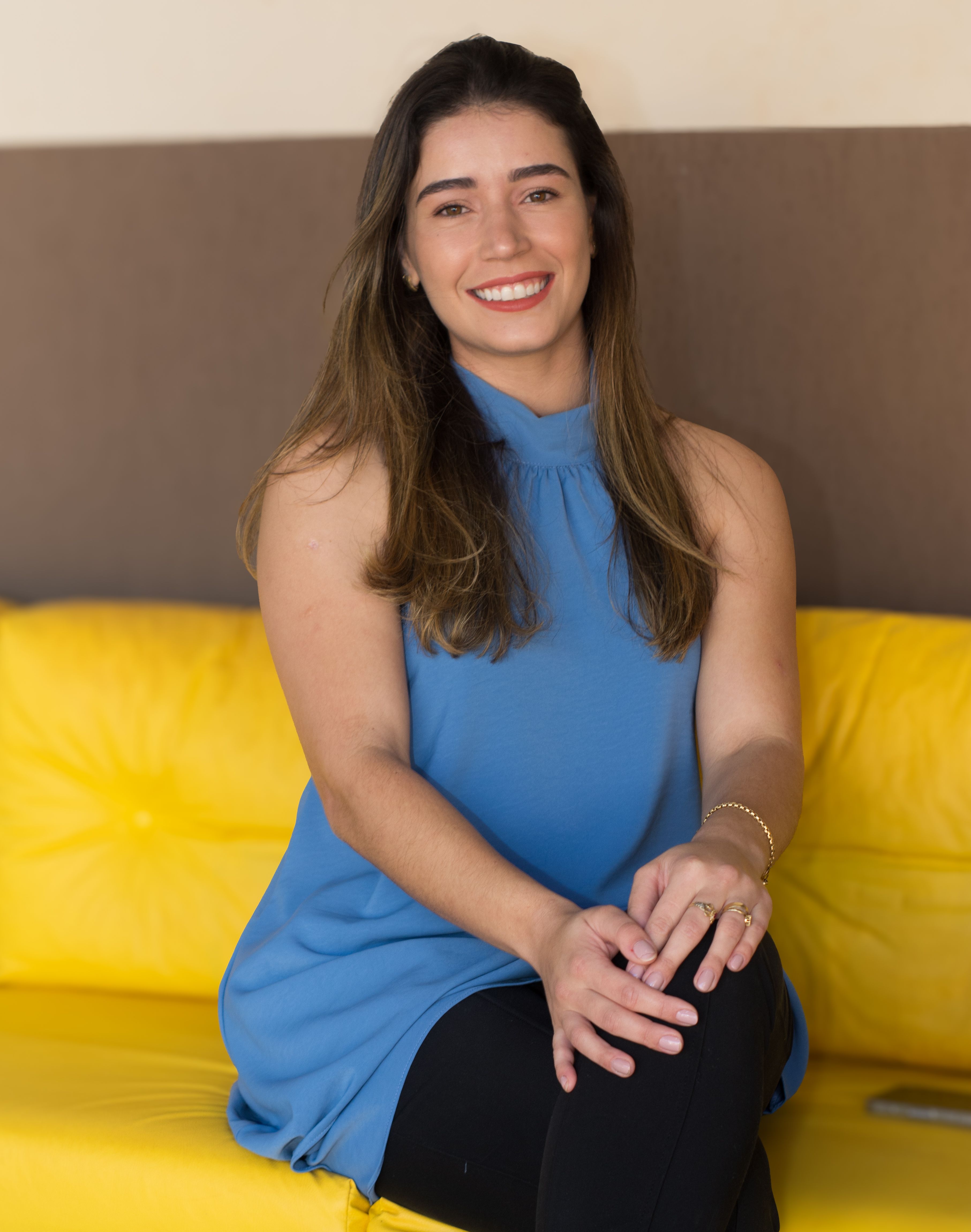 A woman in a blue top sits smiling on a yellow couch, captured in an indoor setting.