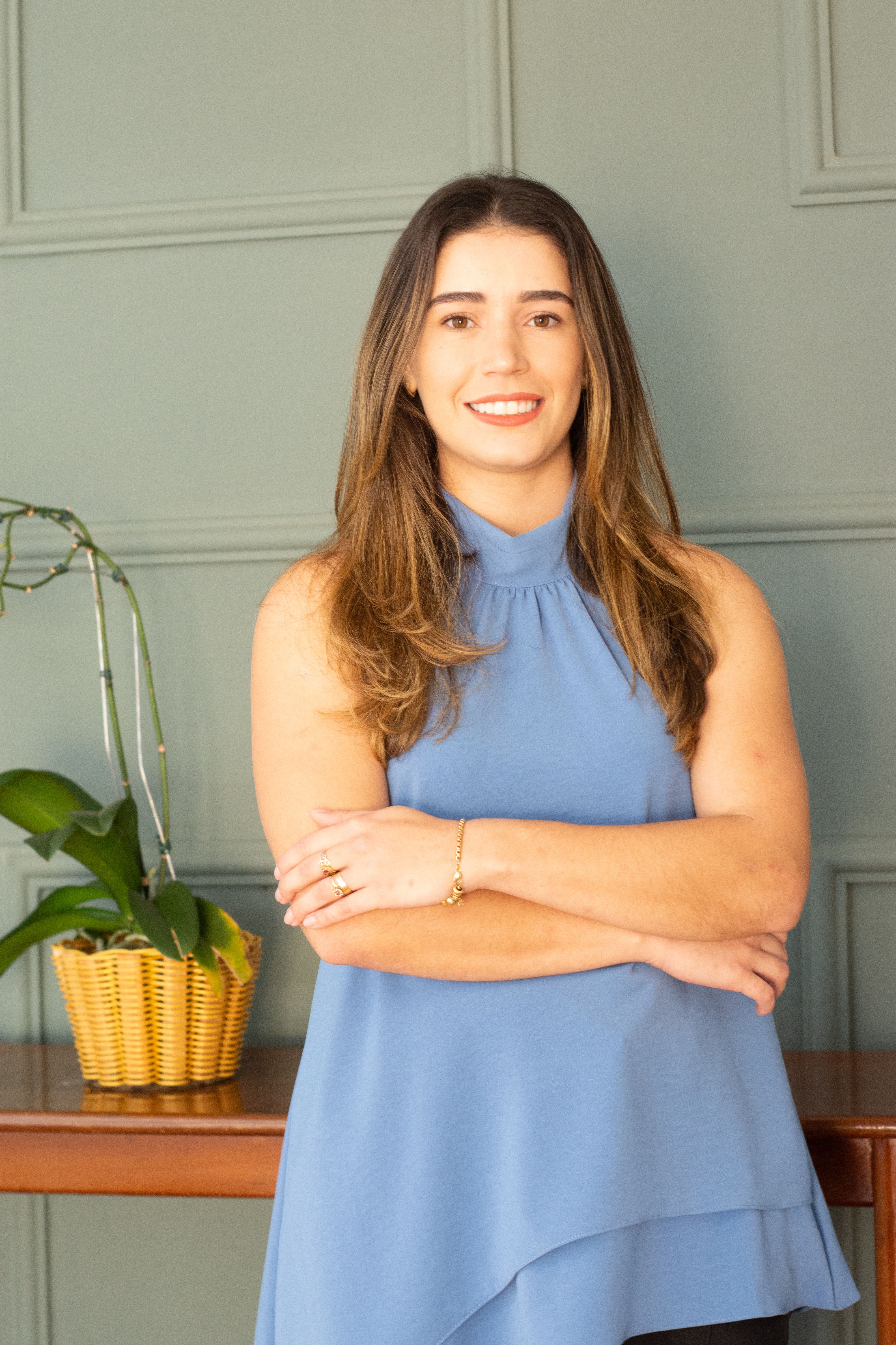 Confident woman standing with arms crossed in an indoor setting beside a plant.