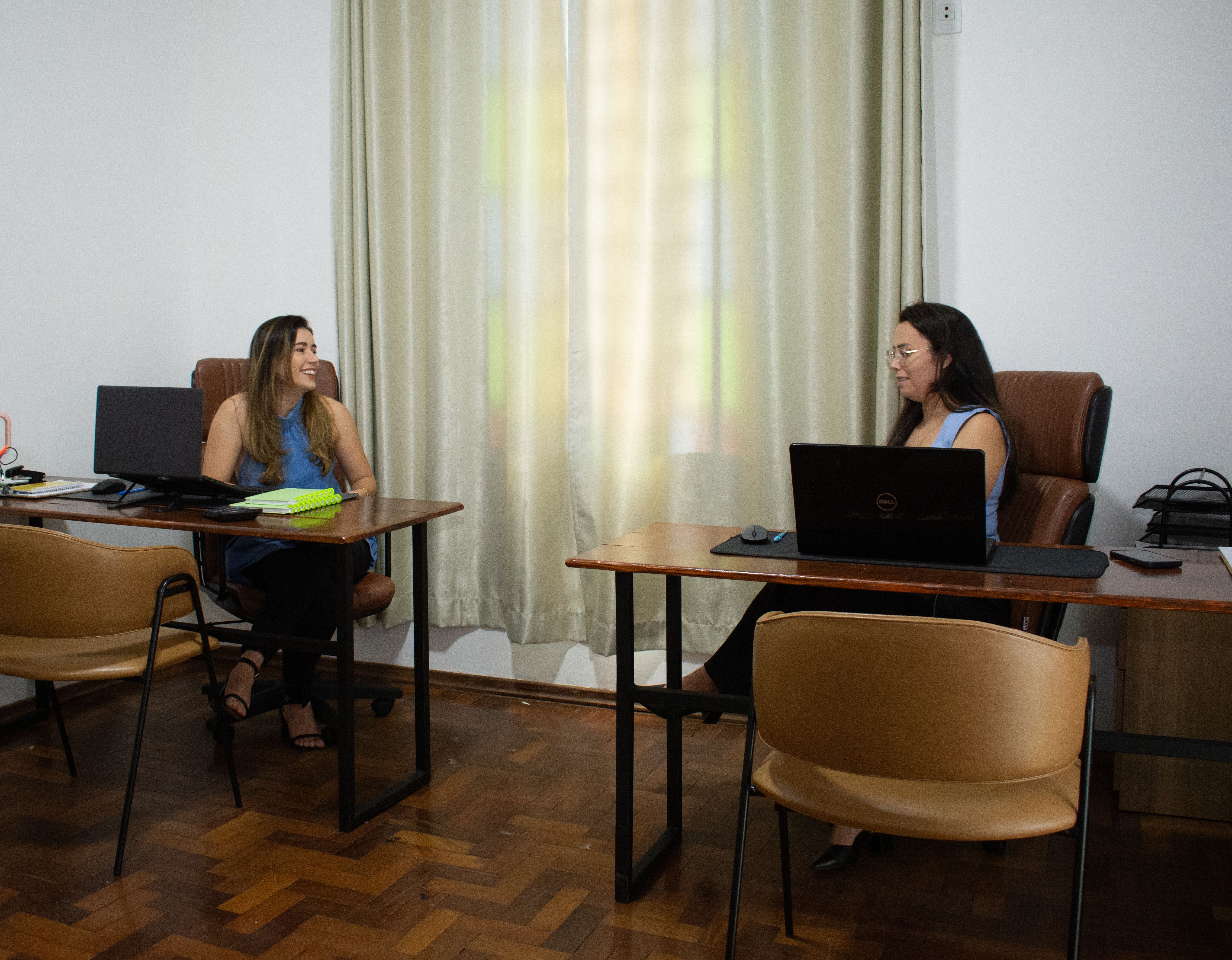 Two businesswomen in an office discussing work across desks with laptops.