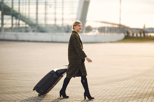 Full length of smiling female traveler in elegant outfit and high heeled boots walking near modern airport building with luggage and looking away