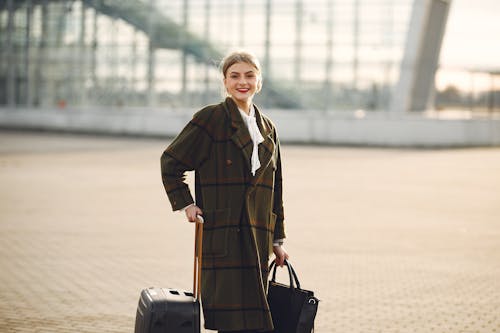 Positive smiling young female traveler in casual warm coat with suitcase and ladies bag standing near airport terminal and looking at camera