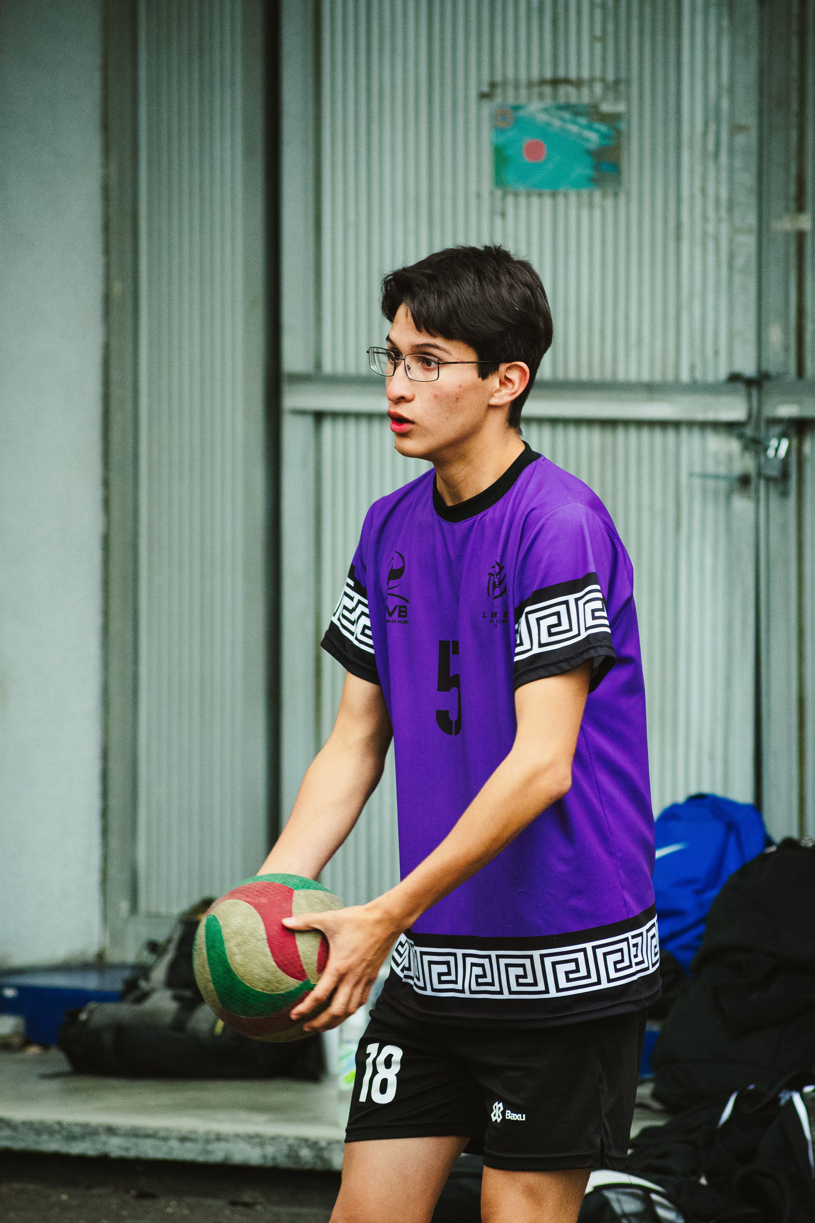 A young male volleyball player in a purple jersey prepares to serve on an urban outdoor court in Mexico City.