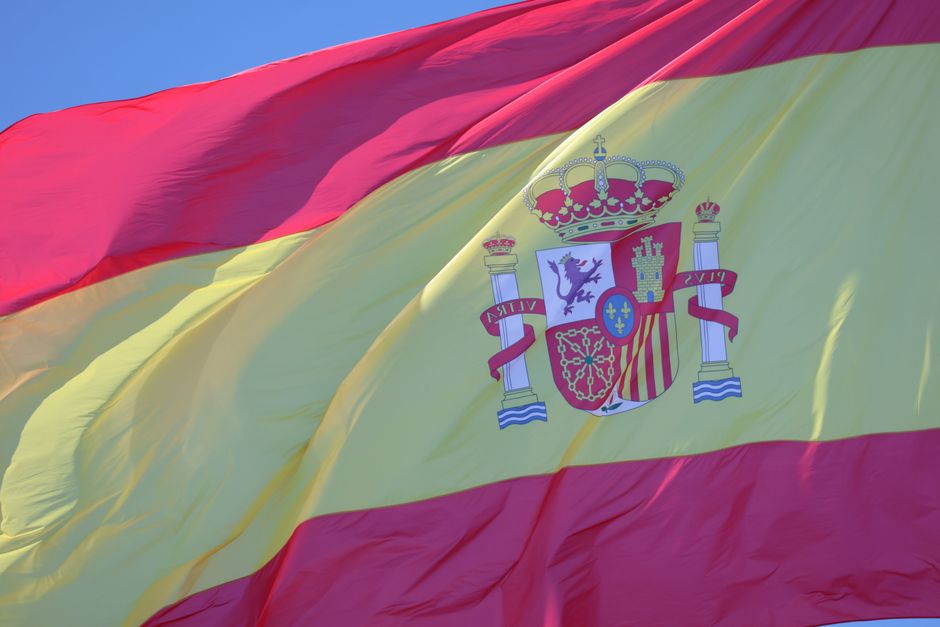 Acercamiento de la bandera de España ondeando contra un cielo azul claro, con colores rojos y amarillos intensos.