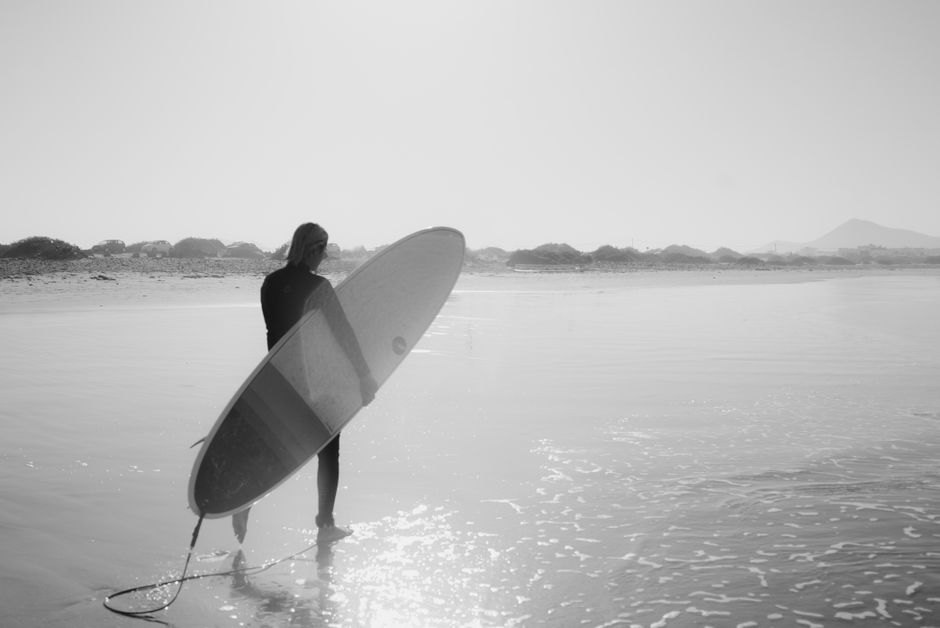 Black and white back view of anonymous guy in wetsuit carrying surfboard tied to leg while going to sea on summer sunny day
