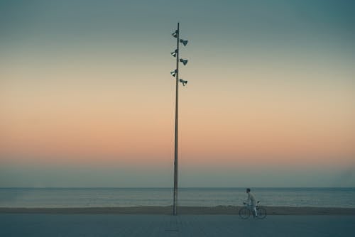 Side view of anonymous person riding on bicycle along seafront near lonely pillar against colorful cloudless sky during sunset