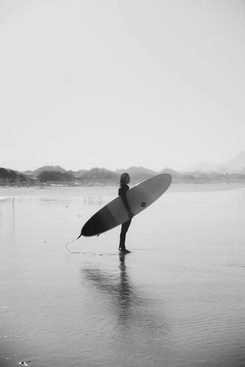 Foto De Escala De Grises Del Hombre Que Sostiene La Tabla De Surf De Pie En La Playa