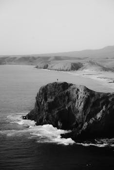 Black and white view of a rugged coastal cliff with crashing waves.