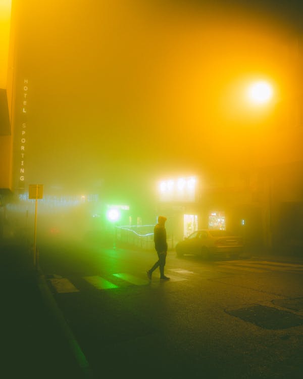 Side view of anonymous person crossing road on crosswalk on illuminated city street covered with fog at night
