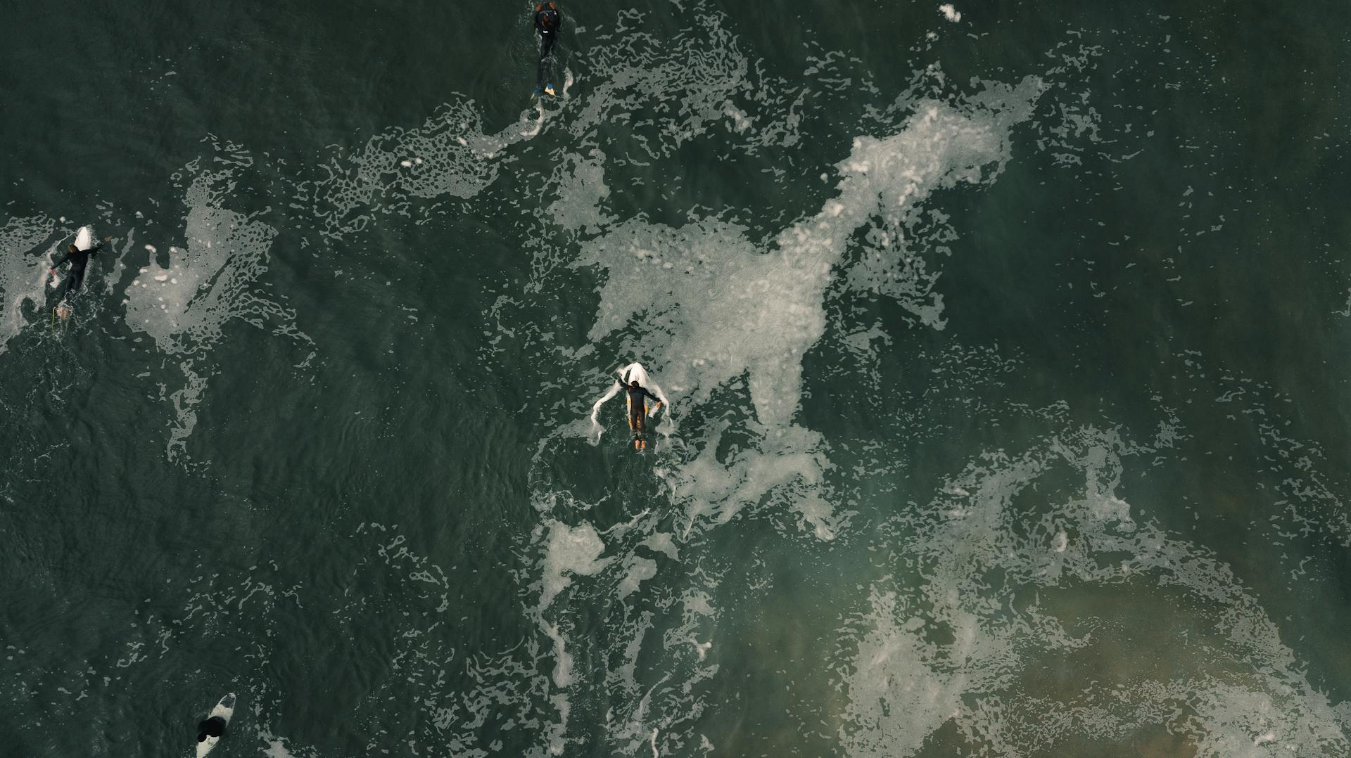 Aerial view of surfers in springsuits rowing surfboards in foamy water waves in ocean