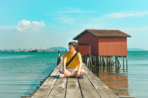 Free A young man sits peacefully on a wooden pier in Penang, Malaysia, next to a red hut over the sea. Stock Photo