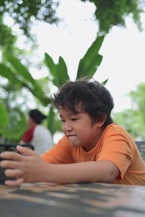 Free Young boy in an orange shirt focuses on smartphone while sitting outdoors among lush greenery. Stock Photo