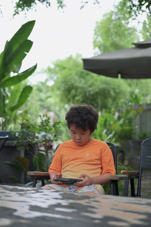 Free Young boy engaged with tablet in a lush garden, surrounded by greenery and outdoor furniture. Stock Photo