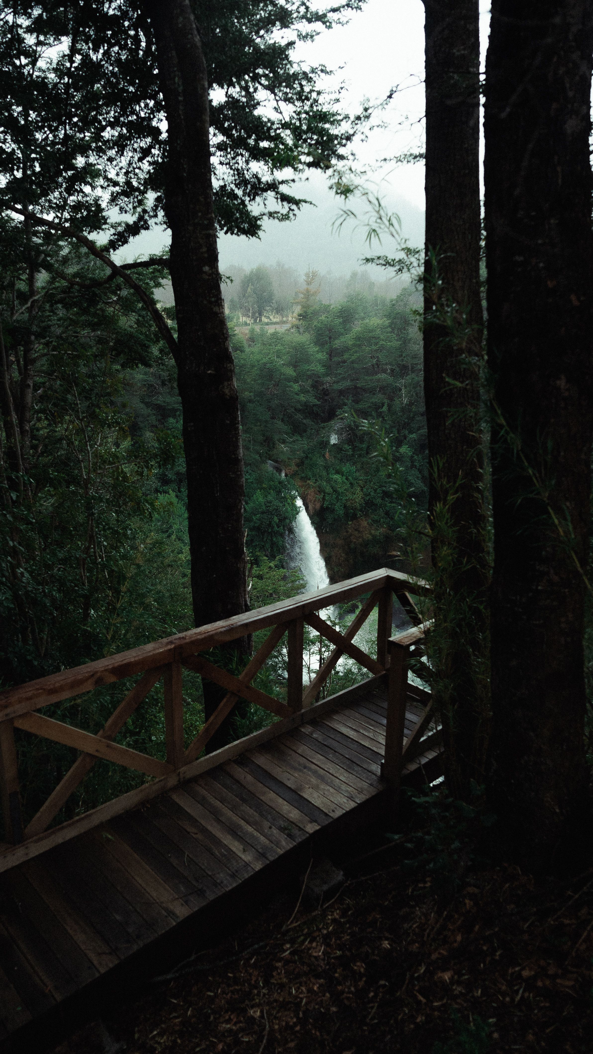 From above of narrow waterfall near rocks and wooden bridge with fence surrounded by green trees in mist in daylight under cloudy sky