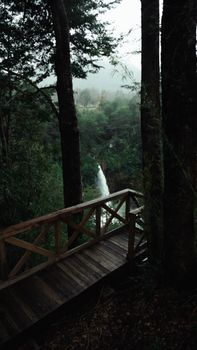 From above of narrow waterfall near rocks and wooden bridge with fence surrounded by green trees in mist in daylight under cloudy sky
